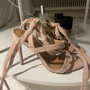 Beiga sandaler med snörning och klack - Snygga beige/rosa sandaler med höga klackar och långa band som snörs runt vristen. Skorna har öppen tå och är gjorda i mocka för en lyxig känsla. Perfekta för att lyfta din outfit med en trendig touch.