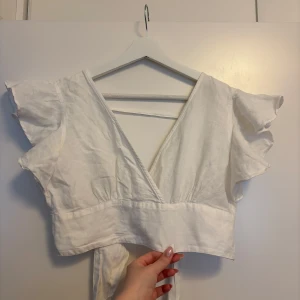 Vit omlottopp med volang från Bik Bok - Superfin vit omlottopp från Bik Bok i linneblandning. Toppen har korta volangärmar, djup v-ringning och knytband i midjan. Perfekt till sommaren och ger en fräsch och luftig känsla. Snyggt croppad modell som passar till både jeans och kjol.