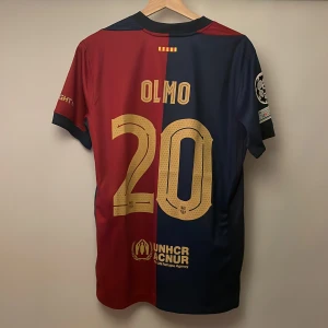 FC Barcelona matchtröja Olmo #20 - Snygg FC Barcelona matchtröja replika med Olmo och nummer 20 på ryggen. Tröjan är röd och marinblå med guldiga detaljer, officiella klubbmärket, Nike-logga och sponsorer som Spotify. Kortärmad modell i lätt funktionsmaterial, Champions League-märke på ärmen. Handtvätt gäller!