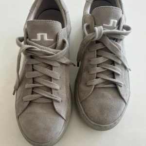 Grå mockasneakers från J.Lindeberg - Snygga sneakers från J.Lindeberg i ljusgrå mocka med matchande grå sula. Skorna har klassisk rund tå, platt sula och snörning framtill. Diskret J.Lindeberg-logga på plösen. Perfekta för en clean och stilren look. Använda fåtal ggr, mycket gott skick! 