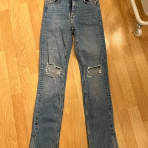 Blå jeans från Zara - Snygga blå jeans från Zara med slitna hål på båda knäna. Modellen är hög midja och klassisk femficksdesign. Jeansen har dragkedja och knapp framtill och är tillverkade i bomull med lite stretch för skön passform🩷