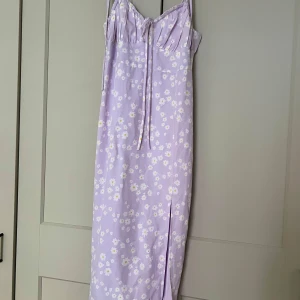 Ljuslila blommig långklänning med slits - Superfin långklänning i ljuslila med vita och gula blommor. Klänningen har smala axelband som knyts, söt snörning vid bysten och en snygg slits framtill. 💜
