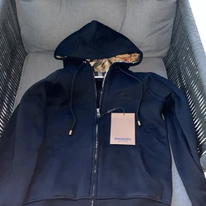 Svart Burberry hoodie med dragkedja - Svart hoodie från Burberry med klassiskt rutigt foder i huvan. Jackan har dragkedja framtill, snörning i huvan och ribbade muddar. Ikonisk Burberry-logga broderad på bröstet och snygga metalldetaljer på snörena. Perfekt för dig som vill ha en stilren och lyxig look.