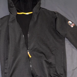Svart zip up från Parajumpers - Svart vindjacka från Parajumpers med huva och dragkedja med orange detalj. Jackan har en snygg patch på ärmen med PJS-logga och texten Anchorage. Perfekt för dig som gillar stilren och sportig look.