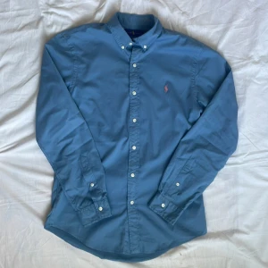 Blå skjorta från Ralph Lauren - Snygg blå skjorta från Ralph Lauren med klassisk button-down krage och broderad rosa logga på bröstet. Skjortan har lång ärm, vita knappar och är tillverkad i mjuk bomull. Perfekt för dig som gillar stilrena och tidlösa plagg.