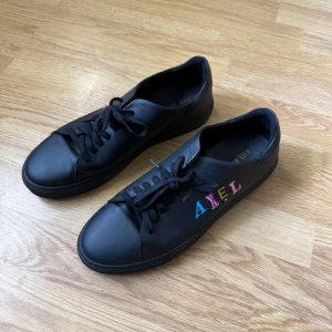 Svarta sneakers Axel Arigato skinn - Svarta sneakers i skinn från Axel Arigato med färgglada bokstäver på sidan. Klassisk rund tå, svarta snören och platt sula. Snygg minimalistisk design med en lekfull touch tack vare de färgade bokstäverna.