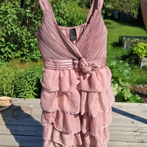 Pale pink volangklänning från VILA - Supersöt rosa kortklänning från VILA med flera lager volanger och v-ringad omlott-topp. Klänningen har breda axelband, markerad midja med rosettdetalj och glansigt tyg som ger en lyxig känsla. Perfekt för dig som vill sticka ut med en romantisk look. Använde den på the eras tour förra året och sen dess har den bara hängt i garderoben. 