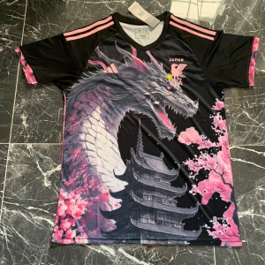 Japan Adidas fotbollströja drake & körsbär - Unik Japan fotbollströja från Adidas med stor drakdesign, körsbärsblommor och japanskt tempel. Svart bas med rosa detaljer och tre ränder på axlarna. Tillverkad i lätt och ventilerande syntetmaterial, perfekt för dig som vill sticka ut på planen.