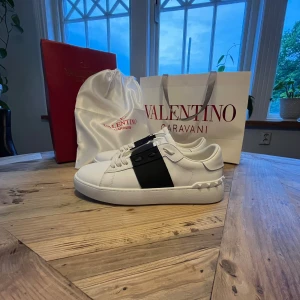 Valentino Garavani Open sneakers vit/svart - Snygga Valentino Garavani Open sneakers i vitt skinn med bred svart läderdetalj över vristen och  nitar baktill. Kommer med originalpåse och låda – perfekt för dig som gillar lyxiga detaljer. Och lådan är lite skadad på sidorna men det är inte så stort, men annars kommer det med lådan kort som satt på de när de var helt nya.