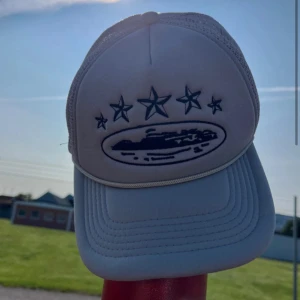 Grå Corteiz keps med broderade stjärnor - Snygg grå keps från Corteiz med broderade svarta stjärnor och motiv framtill. Kepsen har mesh på sidorna och bak för extra ventilation samt justerbar passform. Perfekt accessoar för en streetwear-look.