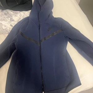 Blå Nike hoodiejacka med dragkedja S - Snygg blå hoodiejacka från Nike med hel dragkedja och svart Nike-logga på bröstet. Jackan har en sportig look med svarta detaljer över bröstet och huva. Perfekt för dig som gillar streetwear och vill ha en bekväm jacka till vardags.