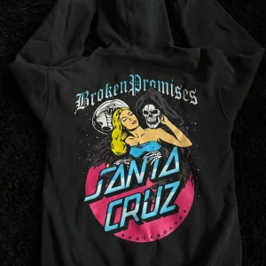 Santa Cruz x Broken Promises hoodie M - Svart hoodie från Santa Cruz x Broken Promises med stor färgglad print på ryggen och mindre tryck på bröstet. Klassisk huva med snörning och känguruficka. Materialet är mjukt och skönt, perfekt för skate eller chill. Grym streetwear-vibe!