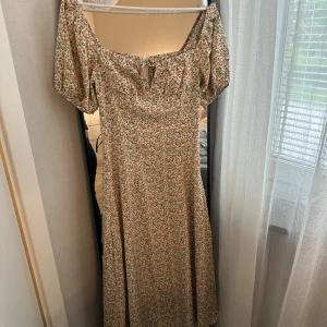 Blommig långklänning med puffärm - Romantisk långklänning med småblommigt mönster i beige, grönt och rosa. Klänningen har offshoulder-modell med puffiga kortärmar och söt knytning framtill. Perfekt för sommarens alla tillfällen.