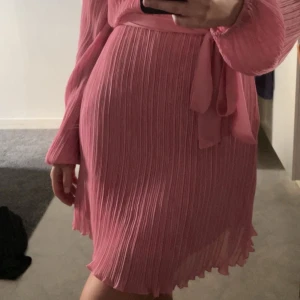 Plisserad rosa klänning från Nelly - Superfin plisserad rosa klänning från Nelly med lång ärm och knytband i midjan. Klänningen har rund halsringning, lätt transparent tyg och vågig kant nertill. Perfekt för dig som gillar en feminin och trendig look.
