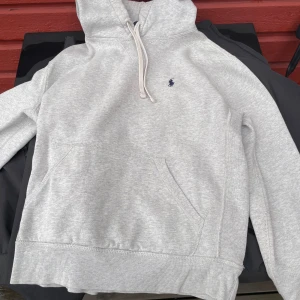 Grå hoodie från Polo Ralph Lauren - Snygg ljusgrå hoodie från Polo Ralph Lauren med klassisk broderad logga i mörkblått på bröstet. Tröjan har huva med snörning, känguruficka och är tillverkad i mjuk bomullsmix. Perfekt för chill dagar eller när du vill ha en clean look. Storlek small men passar xs. Pris kan diskuteras
