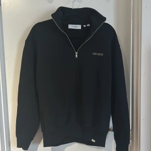 Svart half zip sweatshirt Les Deux - Svart sweatshirt från Les Deux med half zip-dragkedja och broderad logga på bröstet. Tröjan har hög krage och lång ärm, perfekt för en clean och stilren look. Tillverkad i mjuk bomull för maximal komfort.