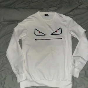 Fendi tröja  - Vit långärmad tröja från Fendi med ett coolt ansikte tryckt på bröstet. Ögonen är svarta med röda detaljer och munnen är en dragkedja. Tröjan har ribbade muddar och är gjord i mjuk bomull. Perfekt för dig som vill sticka ut med en unik look.