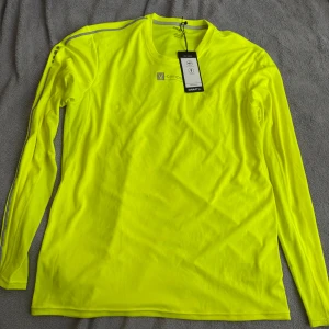 Gul långärmad träningströja från Craft - Neongul långärmad träningströja från Craft med rund hals och diskreta reflexdetaljer på axeln. Tillverkad i lätt och ventilerande polyester, perfekt för löpning eller annan träning. Snygg logga framtill och sportig passform.