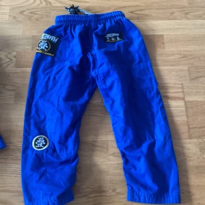 Blåa Tatami Jiu Jitsu byxor M0 - Säljer ett par blåa Jiu Jitsu byxor från Tatami i storlek M0. Byxorna har resår och snörning i midjan, broderade patchar och är tillverkade i 100% bomull. Perfekta för träning och kampsport.