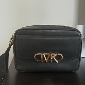 Svart axelväska från Michael Kors - Stilren svart axelväska från Michael Kors i skinn med guldfärgade detaljer och MK-logga framtill. Väskan har en praktisk dragkedja upptill och ett extra fack på framsidan. Perfekt storlek för det viktigaste och enkel att matcha till olika outfits.