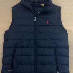 Mörkblå väst från Polo Ralph Lauren - Snygg mörkblå dunväst från Polo Ralph Lauren med klassisk quiltad design och dragkedja framtill. Västen har två fickor med dragkedja och ikonisk röd logga broderad på bröstet. Perfekt att slänga över en hoodie eller tröja för extra värme. 