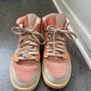 Nike Air Jordan 1 Mid Peach EUR 40 - Nike Air Jordan 1 Mid i snygga persikofärgade och beige toner. Skorna har en mix av skinn och mocka, med klassisk snörning och ikoniska Jordan-detaljer. Perfekt för dig som vill sticka ut med en unik färgkombo och streetstyle-vibe.