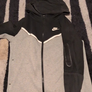 Nike svart och grå hoodiejacka xs - Nike hoodiejacka i svart och grått med vit logga på bröstet. Jackan har hel dragkedja, huva och en snygg vit detalj över bröstet. Tillverkad i mjuk bomullsblandning, perfekt för chill dagar eller när du vill ha en sportig look. Den är lite sol bränt 