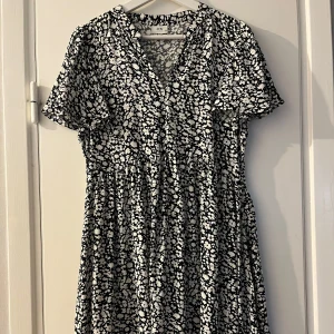 Svartvit blommig klänning från JDY - Supersöt svartvit kortklänning från JDY med blommigt mönster, v-ringning och korta ärmar. Klänningen har en avslappnad passform och är gjord i ett lätt material som känns skönt mot huden. Perfekt för dig som gillar en chill och trendig stil.