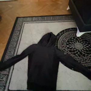 Svart hoodie från Calvin Klein Jeans - Svart hoodie från Calvin Klein Jeans med klassisk huva och stort CK-logga på ärmen. Tröjan har lång ärm och en clean, stilren look som passar perfekt till streetwear. Mjuk och skön, perfekt för lager-på-lager under kyliga dagar.