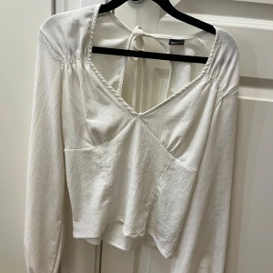 Vit omlottopp med knyt från Gina Tricot - Supersnygg vit omlottopp från Gina Tricot i storlek L. Toppen har lång ärm, v-ringning och knyt i ryggen som ger en öppen rygg-effekt. Materialet är lätt strukturerat och känns luftigt. Perfekt att styla med jeans eller kjol för en trendig look. Använd 1 gång 