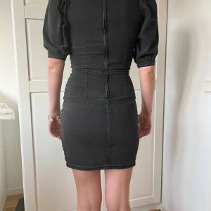 Svart tight kort klänning med puffärm - Snygg svart kort klänning i jeansmaterial med markerad midja och puffiga korta ärmar. Klänningen har en tight passform och synliga sömmar som ger en cool look. Perfekt för dig som gillar en stilren men edgy vibe.