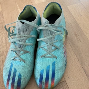 Turkosa fotbollsskor med regnbågsdetaljer - Säljer ett par lätta turkosa Adidas fotbollsskor med snörning och färgglada regnbågsdetaljer på sidorna. Skorna har plös i mesh, rund tå och dobbar för bra grepp på planen. Perfekta för dig som vill sticka ut på matchen!