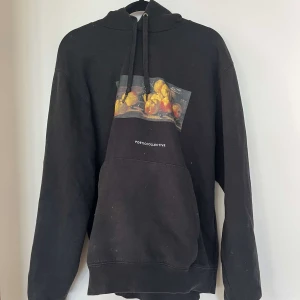 Svart hoodie Poetic Collective M - Svart hoodie från Poetic Collective med konstnärligt fruktmotiv på bröstet och vit logga. Klassisk känguruficka, huva med snörning och mjukt material. Perfekt för dig som gillar streetwear med en artsy twist.