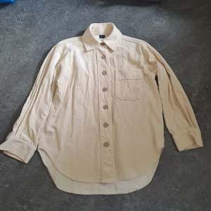 Beige manchester overshirt från Gina Tricot - Snygg beige overshirt i manchester från Gina Tricot. Skjortan har klassisk krage, bröstficka och stora knappar framtill. Långärmad modell med avslappnad passform, perfekt att styla lager på lager.