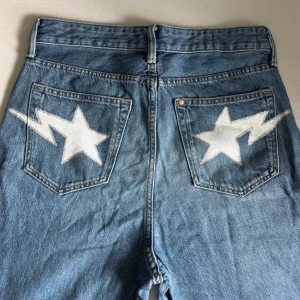 Loose fit jeans med stjärn-detalj H&M - Blå loose fit jeans från H&M med coola vita stjärnor och blixtar på bakfickorna som jag målat själv med textilfärg. Klassisk femficksmodell i denim med rak passform och normal midja. Perfekta för dig som gillar en avslappnad och unik stil.