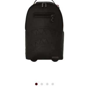 Svart ryggsäck från Sprayground - Limited edition svart ryggsäck från Sprayground med ikoniskt hajtandsmönster i relief på framsidan. Tillverkad i skinnimitation med dragkedja framtill och rutiga detaljer på axelremmarna. Rymligt huvudfack och innerficka med röd etikett.