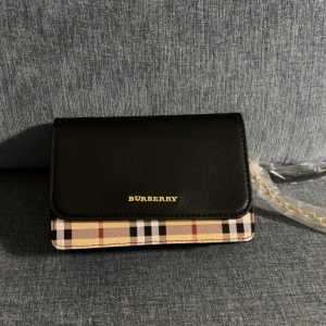 Burberry rutig axelväska med kedja - Helt ny axelväska från Burberry med klassiskt rutigt mönster i beige, svart, vitt och rött. Väskan har svart lock i skinn och guldfärgad kedjerem. Perfekt storlek för det viktigaste och stängs med magnetknapp. Ikonisk och stilren design. 