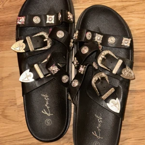 Svarta sandaler med silverspännen - Coola svarta sandaler från Krush med flera remmar dekorerade med silvriga detaljer och spännen. Snygga metalliska nitar och mönstrade spännen ger en edgy vibe. 