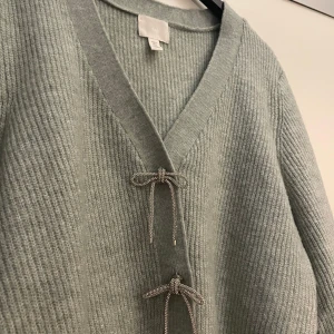 Ljusgrå/mintgrön stickad kofta från H&M - Superfin ljusgrå stickad kofta från H&M med v-ringning och två snygga knappar framtill. Koftan är ribbstickad, har lång ärm och en croppad passform som ger en modern vibe. Perfekt att slänga över en topp för en chill look.