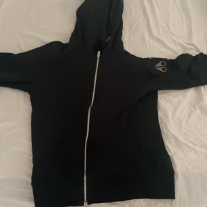 Svart hoodie från Moose Knuckles - Svart hoodie från Moose Knuckles med silverfärgad dragkedja och ikonisk logga i metall på ärmen. Tröjan har huva och långa ärmar, perfekt för en chill och stilren look. Tillverkad i mjuk bomullsmix som känns skön mot huden. Finns en pytteliten skavank som knapps syns. Nypris 3200.