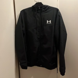 Svart vindjacka från Under Armour - Svart vindjacka från Under Armour med huva och dragkedja framtill. Jackan har en stilren look med logga på bröstet och justerbar snörning i huvan. Perfekt för blåsiga dagar och sportiga outfits.
