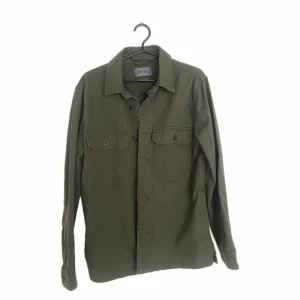 Olivgrön overshirt från Primark - Stilren olivgrön overshirt från Primark med klassisk krage och två bröstfickor med knappar. Skjortan har lång ärm och stängs med knappar framtill. Perfekt att bära öppen över en t-shirt eller stängd för en clean look.