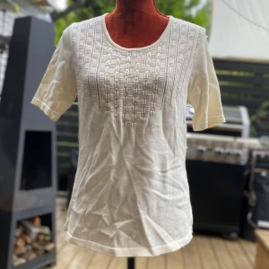 Vit stickad topp från Creation Atelier S - Elegant, beige kortärmad tröja med ett unikt, genombrutet mönster framtill. 100% bomull Längd: 66 cm Bystvidd: 50 cm Katter finns i hemmet!