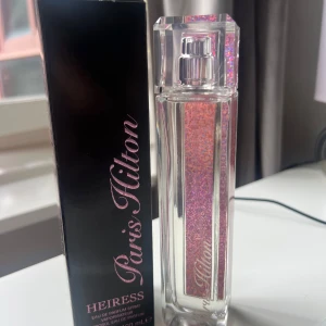 Paris Hilton Heiress Parfym 100ml - Paris Hilton Heiress Eau de Parfum i en snygg, genomskinlig flaska med glittrande rosa insida och silverdetaljer. Flaskan är rektangulär med ett elegant lock och kommer i en svart och rosa förpackning med Paris Hiltons signatur.