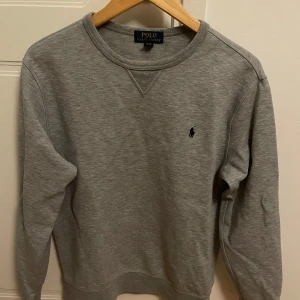 Grå sweatshirt från Polo Ralph Lauren - Klassisk grå sweatshirt från Polo Ralph Lauren med rund halsringning och broderad logga på bröstet. Tröjan har en enkel, stilren design och är tillverkad i mjuk bomullsmix. Perfekt för en avslappnad och snygg look. XS men passar S!