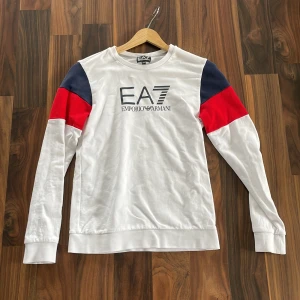 Vit långärmad tröja EA7 Armani - Snygg långärmad tröja från EA7 Emporio Armani i vitt med coola röda och marinblå detaljer på axlarna. Stor logga framtill. Skön passform och tillverkad i mjuk bomull. Perfekt för dig som gillar stilrena och sportiga vibes.
