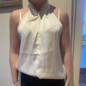 Vit halterneck blus i satin - Säljer en vitt blus i glansig satin med halterneck och snygg knytdetalj vid halsen. Toppen är ärmlös och har en liten knapp i nacken för stängning. Perfekt för dig som vill ha en elegant stil