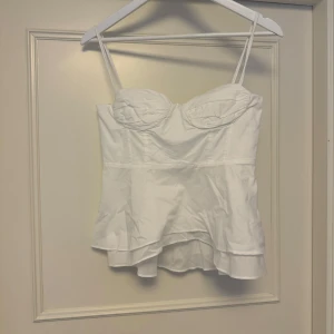 Vit korsett-topp från Zara - Supersnygg vit korsett-topp från Zara med tunna axelband och markerade kupor. Toppen har volangkant nertill och smock baktill för extra komfort. Knytband på ryggen ger en cool detalj. Perfekt att styla med jeans eller kjol för en trendig look. Lappen kvar men testar att sälja här innan jag lämnar tillbaka den!! Storlek S!!