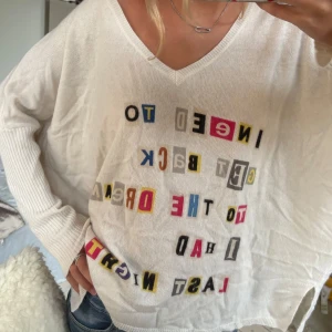 Vit v-ringad tröja med färgglatt tryck - Vit, loose-fit tröja med v-ringning och långa ärmar. Framsidan har ett färgglatt texttryck i olika bokstäver och färger. Ärmarna är ribbstickade och tröjan har en avslappnad passform, perfekt för en chill och trendig look.