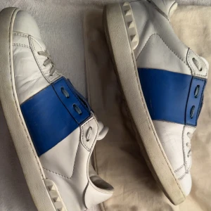 Valentino Garavani sneakers blå/vit - Snygga Valentino Garavani sneakers i vitt skinn med bred blå panel och coola nitar på sulan. Klassisk rund tå, vita snören och robust platt sula. Märkeslogga på plösen och detaljer i blått för en stilren look. Perfekta för dig som gillar exklusiva sneakers.. perfekta inför sommaren och hösten. Man får även med orginal påse. Pris kan diskuteras. Skicket är bra men det finns några skråmor men annars syns det knappt att dom är använda.